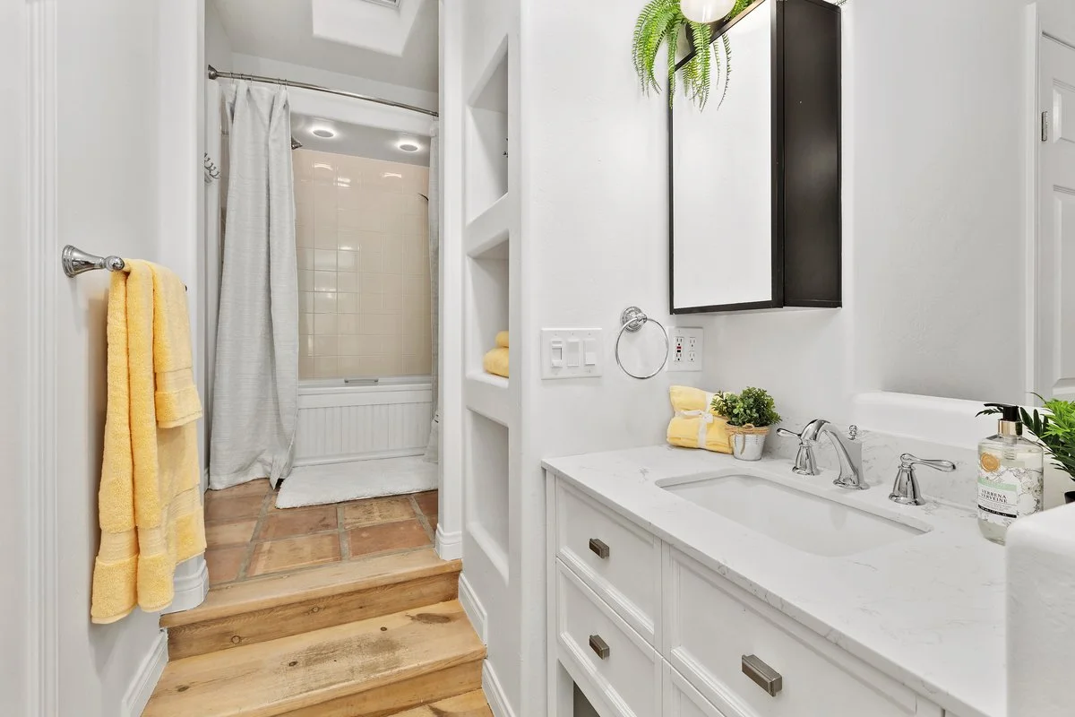 Bathroom with white vanity and black mirror, green plants, yellow towels, and a shower with beige tiles and white curtain