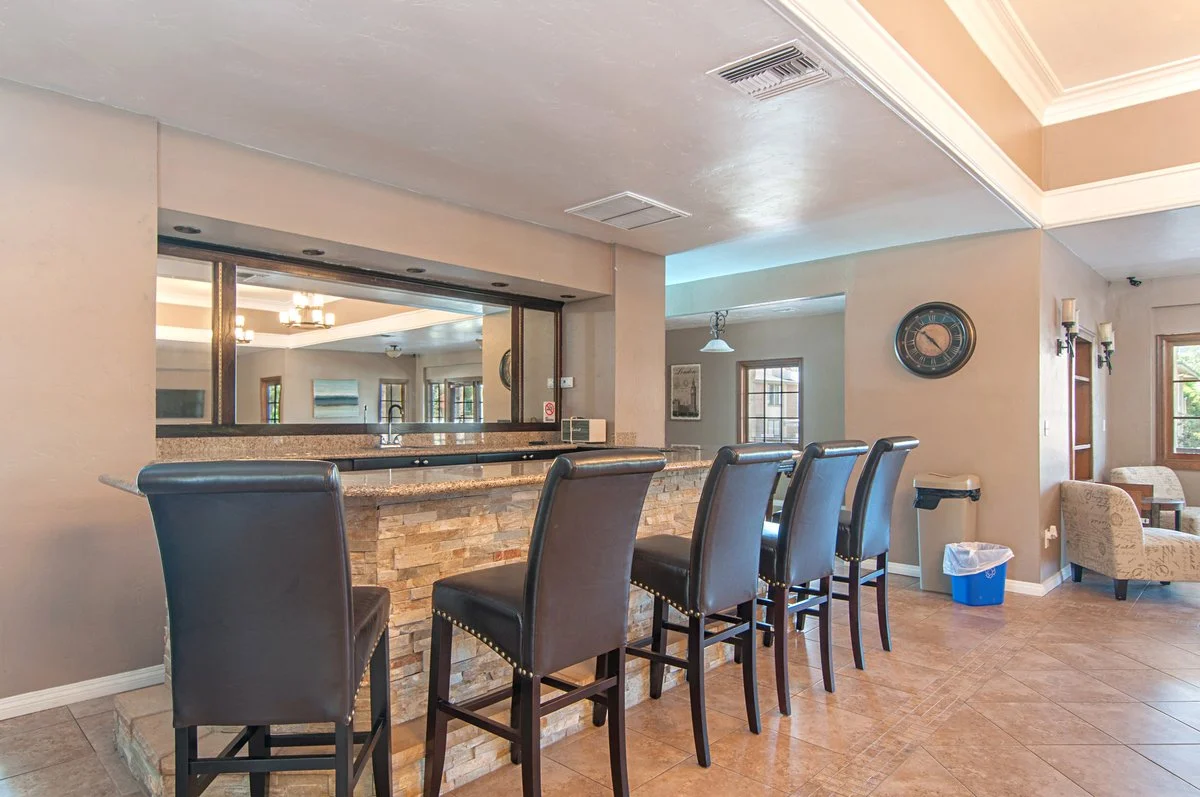 Interior view of a kitchen and sitting area with a bar counter, four black leather chairs, stone facade, open windows, and decorative lighting.