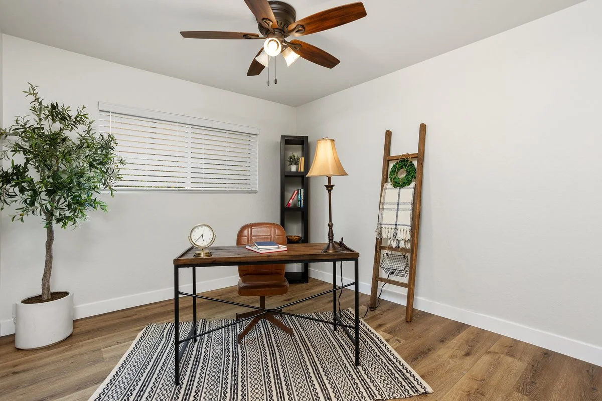An office with a wooden desk, brown leather chair, black shelf, table lamp, clock, ladder with decorative towel, green wreath, potted plant, window with blinds, ceiling fan, and striped rug.