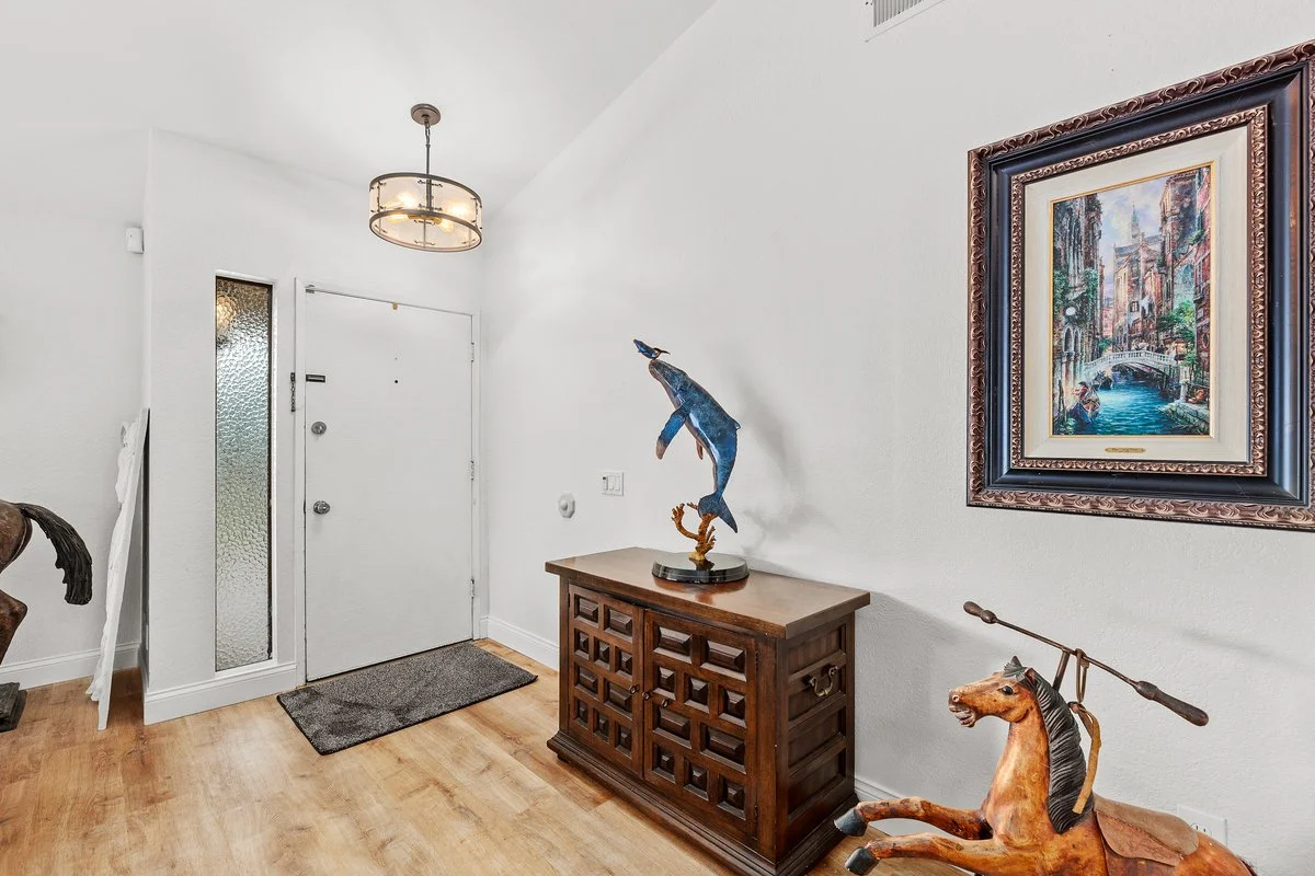 Entryway with white walls, a wooden console table, a framed painting of a European canal scene, a sculpture of a jumping fish on the console, a wooden horse sculpture with a spear through its neck, a modern ceiling light, and a grey welcome mat near 