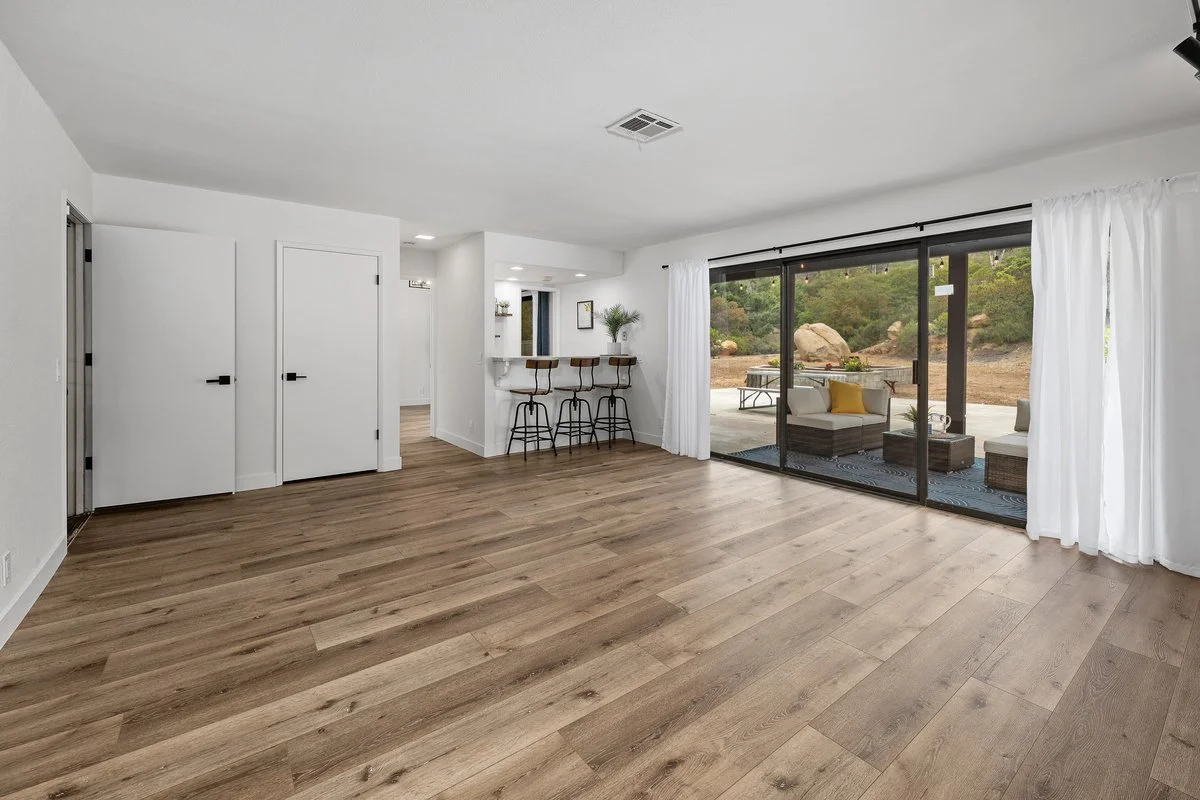 Empty living room with large sliding glass doors leading to outdoor patio with seating, wood flooring, white walls, and curtains.