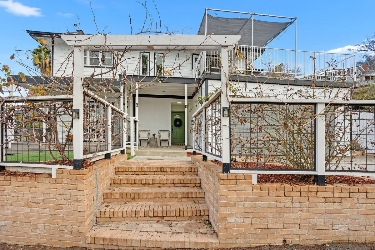 A two-story house with brick steps leading to a front porch, featuring a white railing and a green front door decorated with a wreath. The house has a rooftop terrace with a shade and outdoor seating, and the front yard is enclosed with a white fence