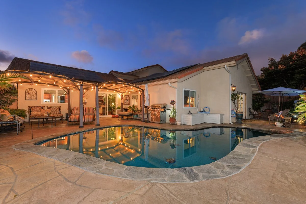 Backyard with illuminated patio, swimming pool, outdoor seating, and a house during dusk