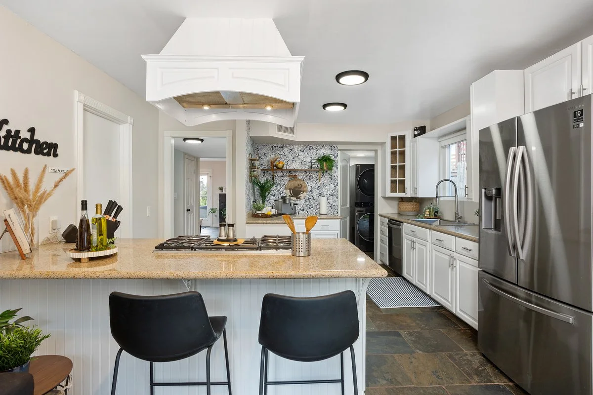 Modern kitchen with white cabinets, a large stainless steel refrigerator, a beige granite island with two black bar stools, and a backsplash with floral pattern tiles.