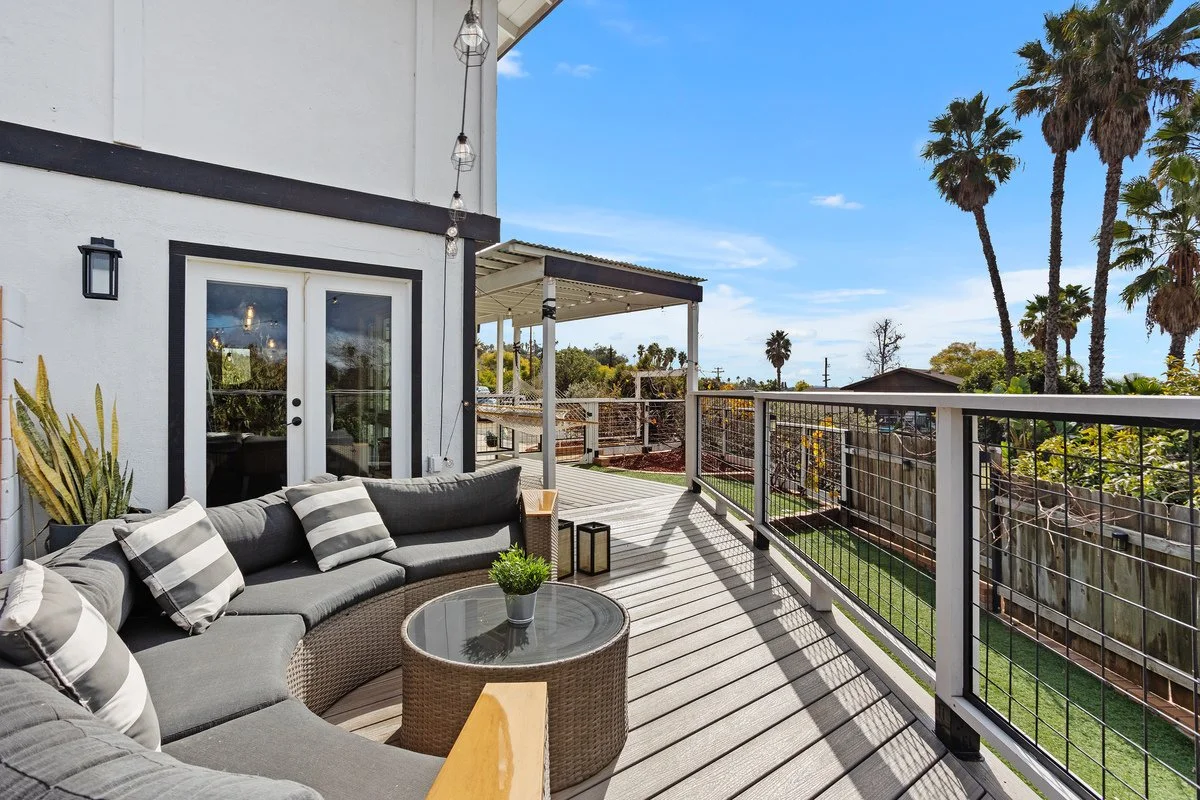 A spacious balcony with a gray sectional sofa with striped pillows, a round wicker coffee table with a small plant, and an outdoor lantern, overlooking a backyard with palm trees and a clear blue sky.
