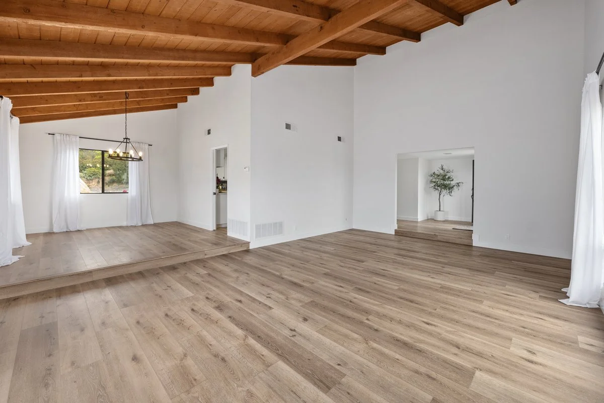 Empty living room with white walls, wood flooring, a wood beamed ceiling, and large windows with white curtains. There is a small elevated section with a chandelier and windows showing greenery outside.