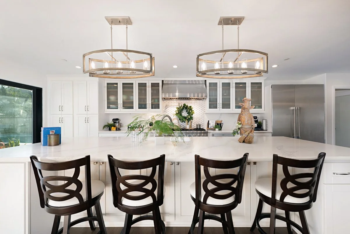 Modern white kitchen with large central island, four black and white chairs, stainless steel appliances, and two circular chandeliers.