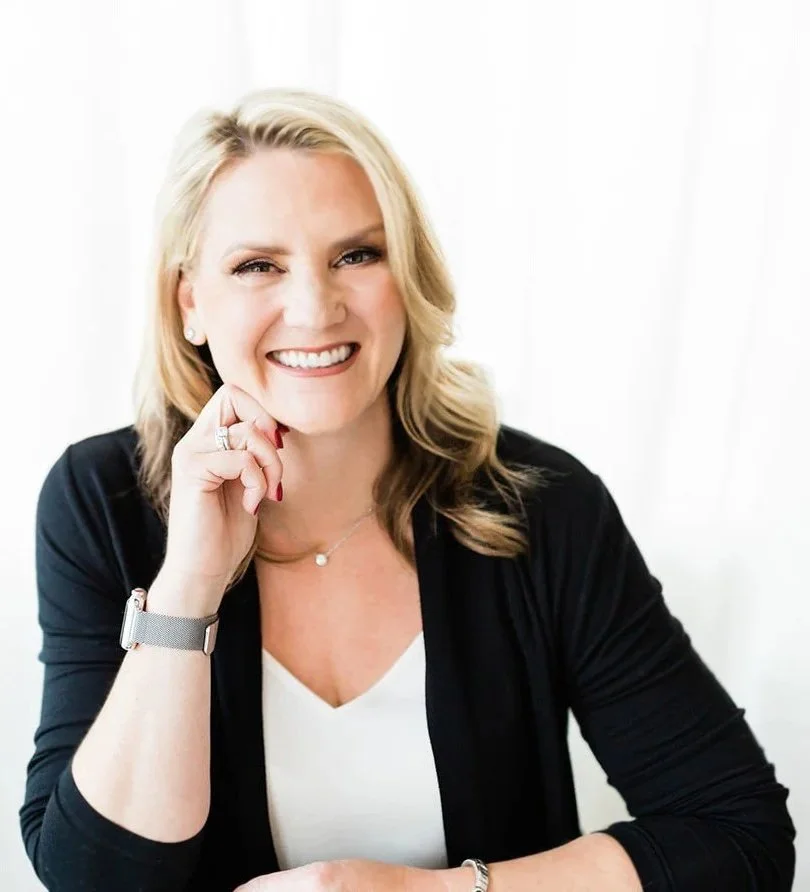 A woman with blonde hair smiling, wearing a black blazer and white top, sitting against a light background.