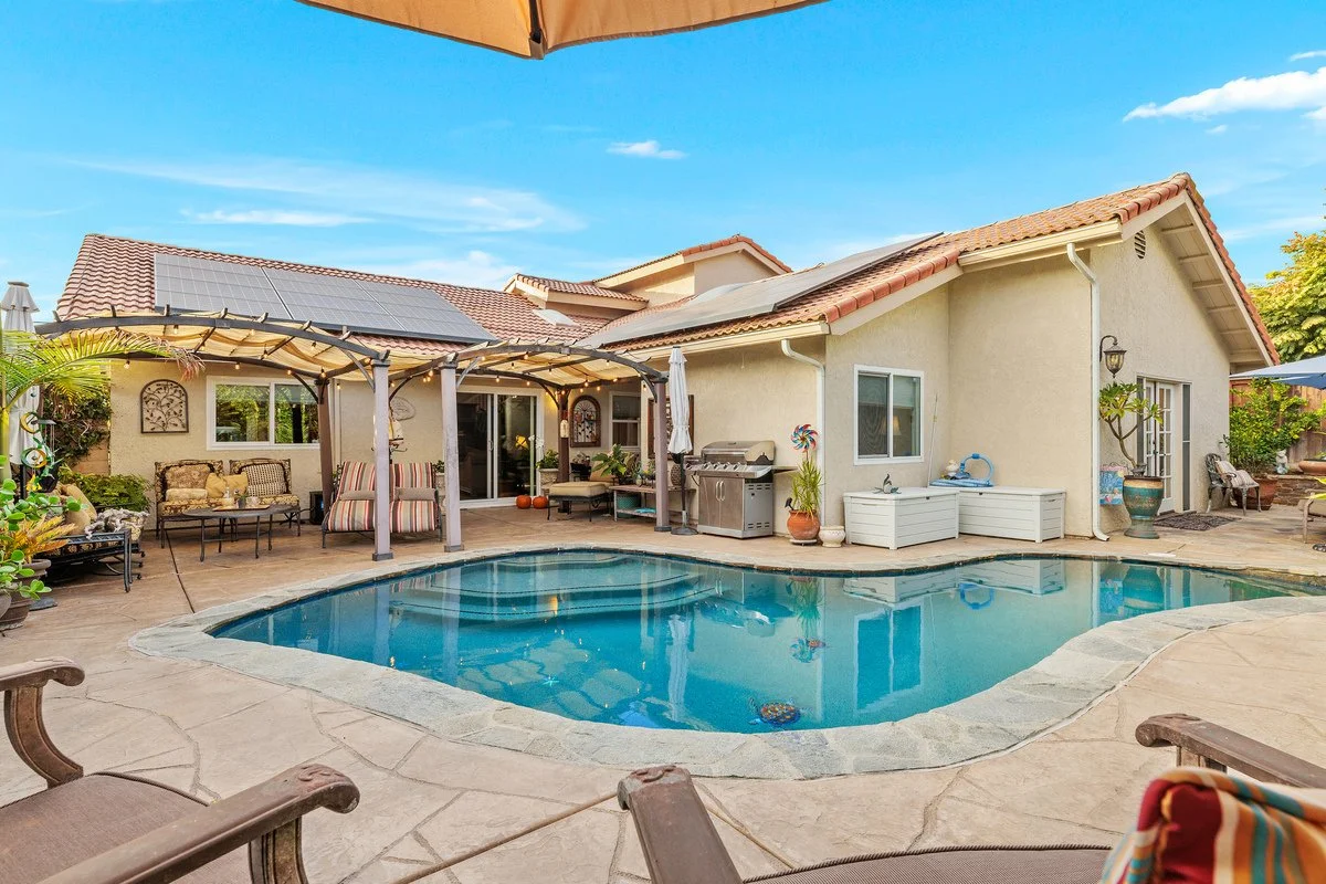 Backyard with a swimming pool, covered patio area with outdoor furniture, barbecue grill, potted plants, and a house with solar panels on the roof. Clear blue sky overhead.