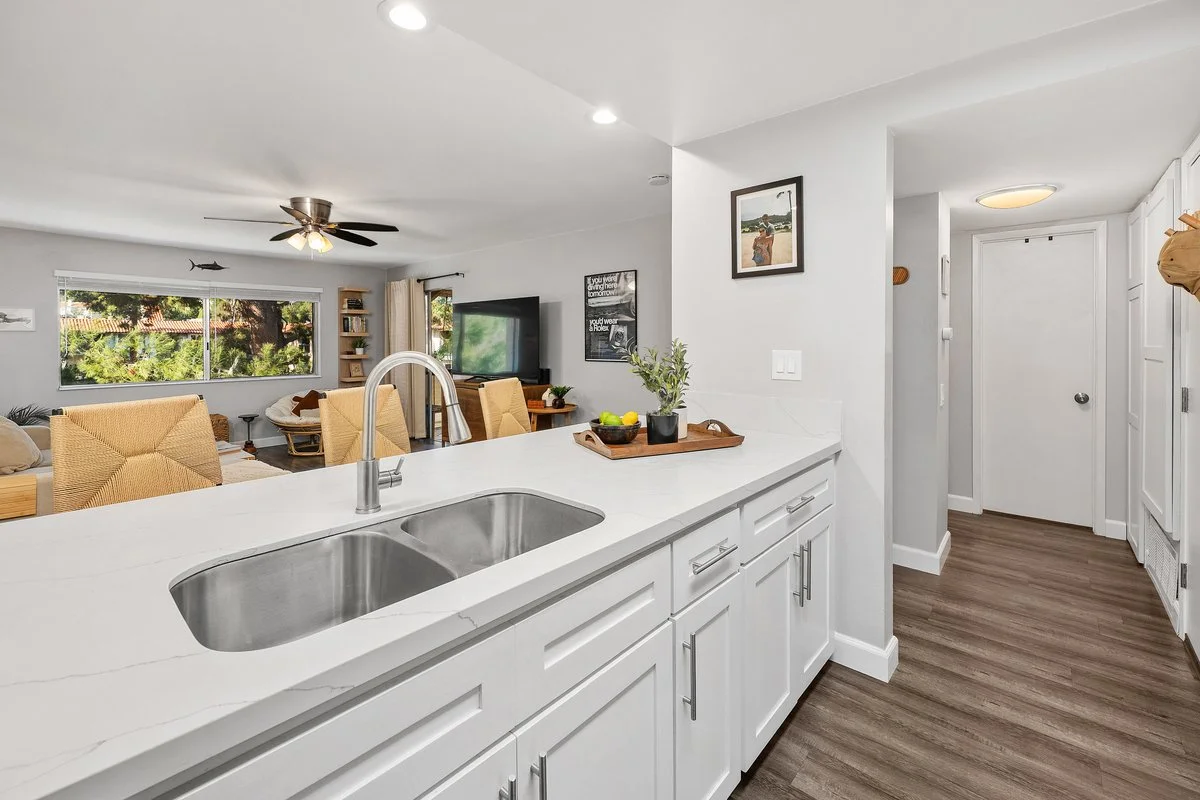 Open kitchen with white cabinets, a double sink, a white countertop, and a small tray with a plant and lemons. In the background, a living room with a television, large window, and beige chairs. Wooden flooring throughout.