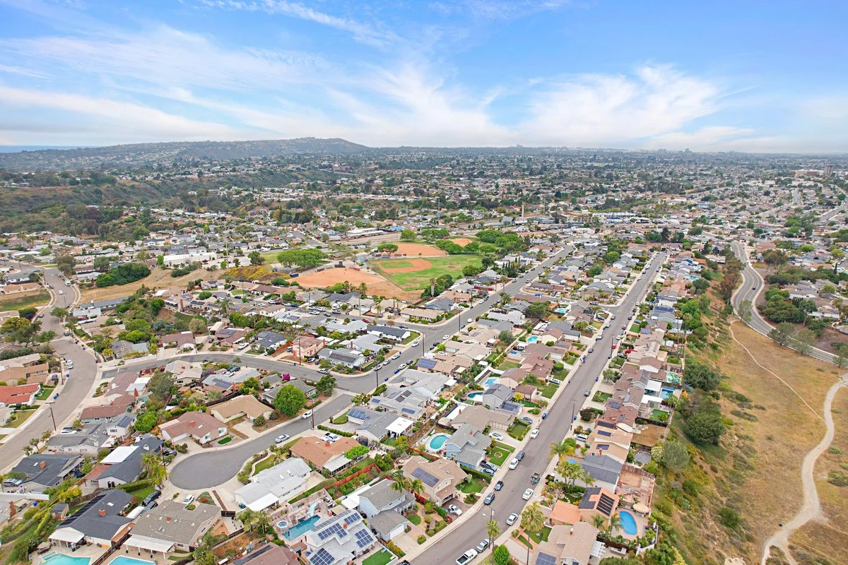 Aerial view of a suburban neighborhood with houses, streets, and a baseball field, with hills and open land in the background under a partly cloudy sky.