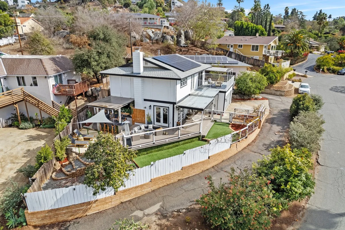 Aerial view of a modern two-story house with solar panels on the roof, a backyard with a lawn and outdoor furniture, surrounded by a white fence and neighboring houses.