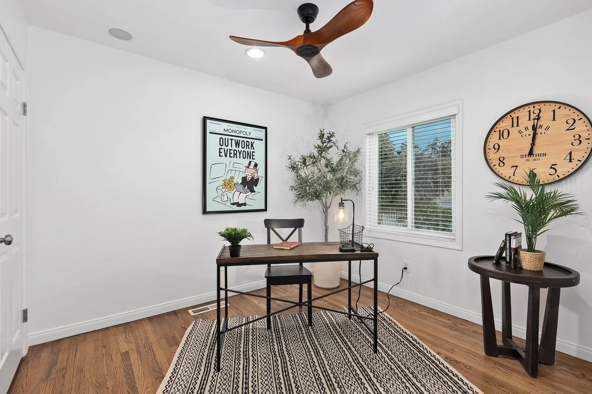Home office with white walls, wooden floor, a wooden desk with a black chair, a wall clock, a potted plant, a window with blinds, a framed Monopoly poster, a table with a plant and books, and a ceiling fan.