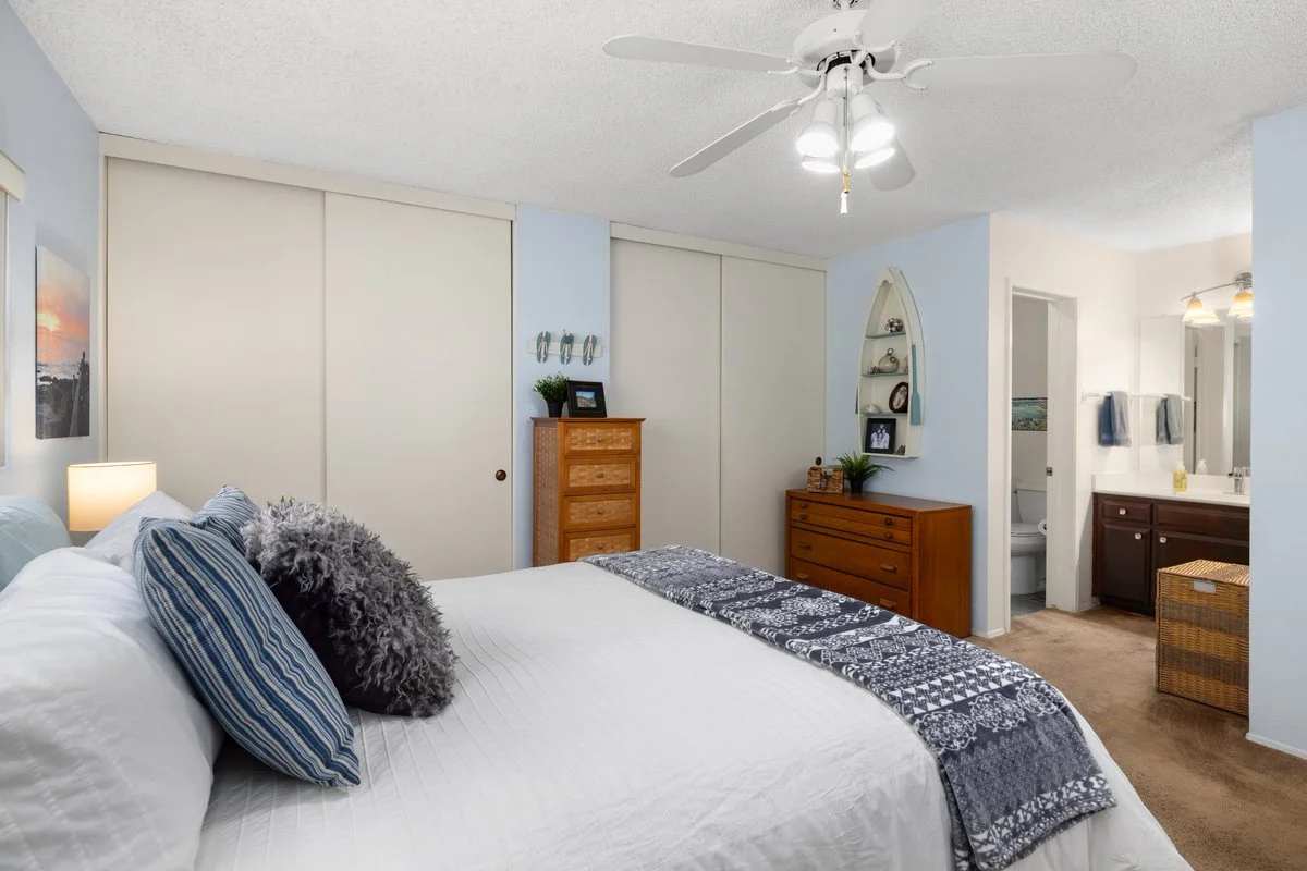 Bedroom with a white bed, blue pillows, and a dark gray decorative pillow, a bedside lamp, a wooden dresser, a floating shelf with decor, and a doorway to an bathroom area.