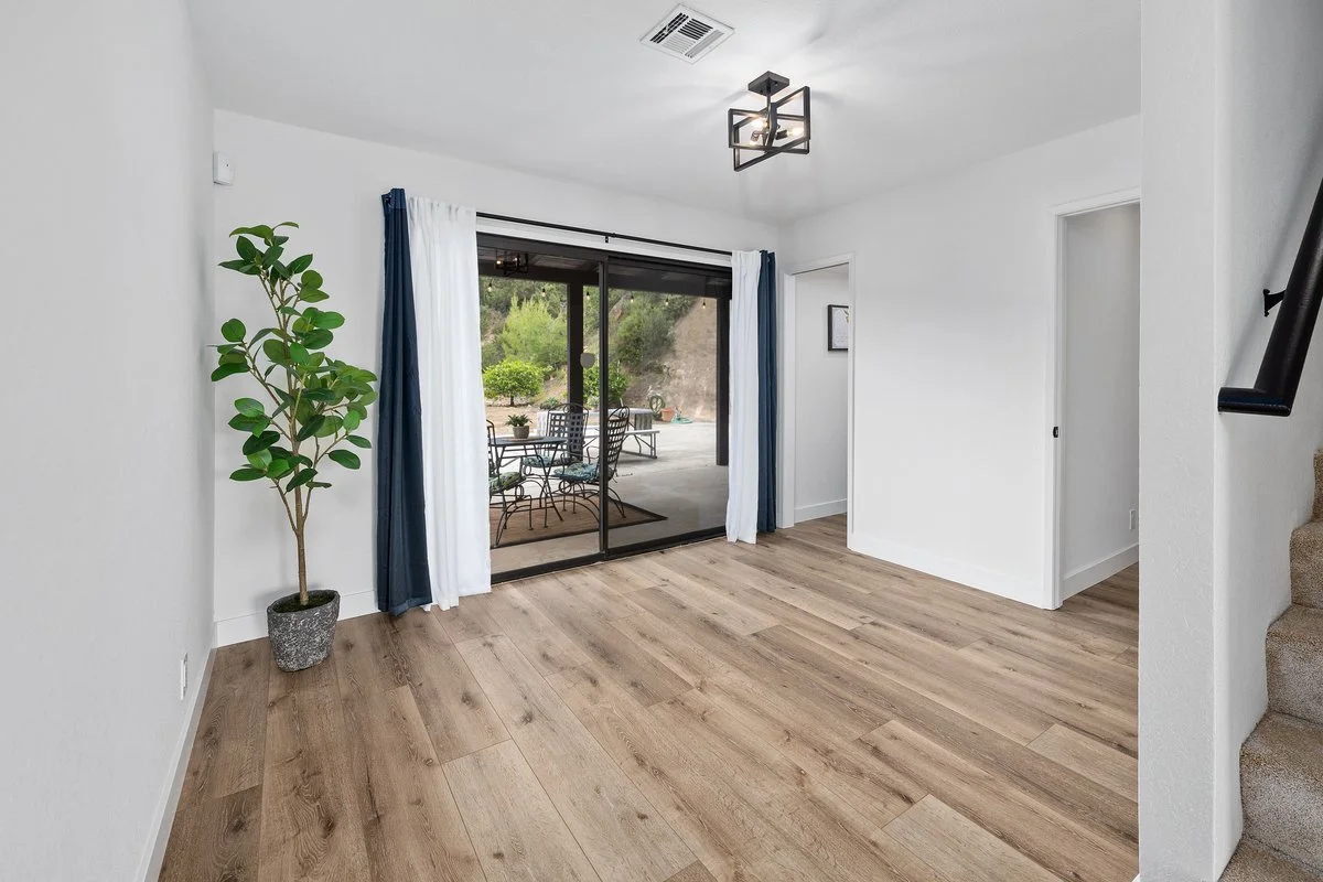 Empty living room with light wood flooring, white walls, sliding glass door with curtains leading to patio, potted plant, modern ceiling light, staircase on right side.