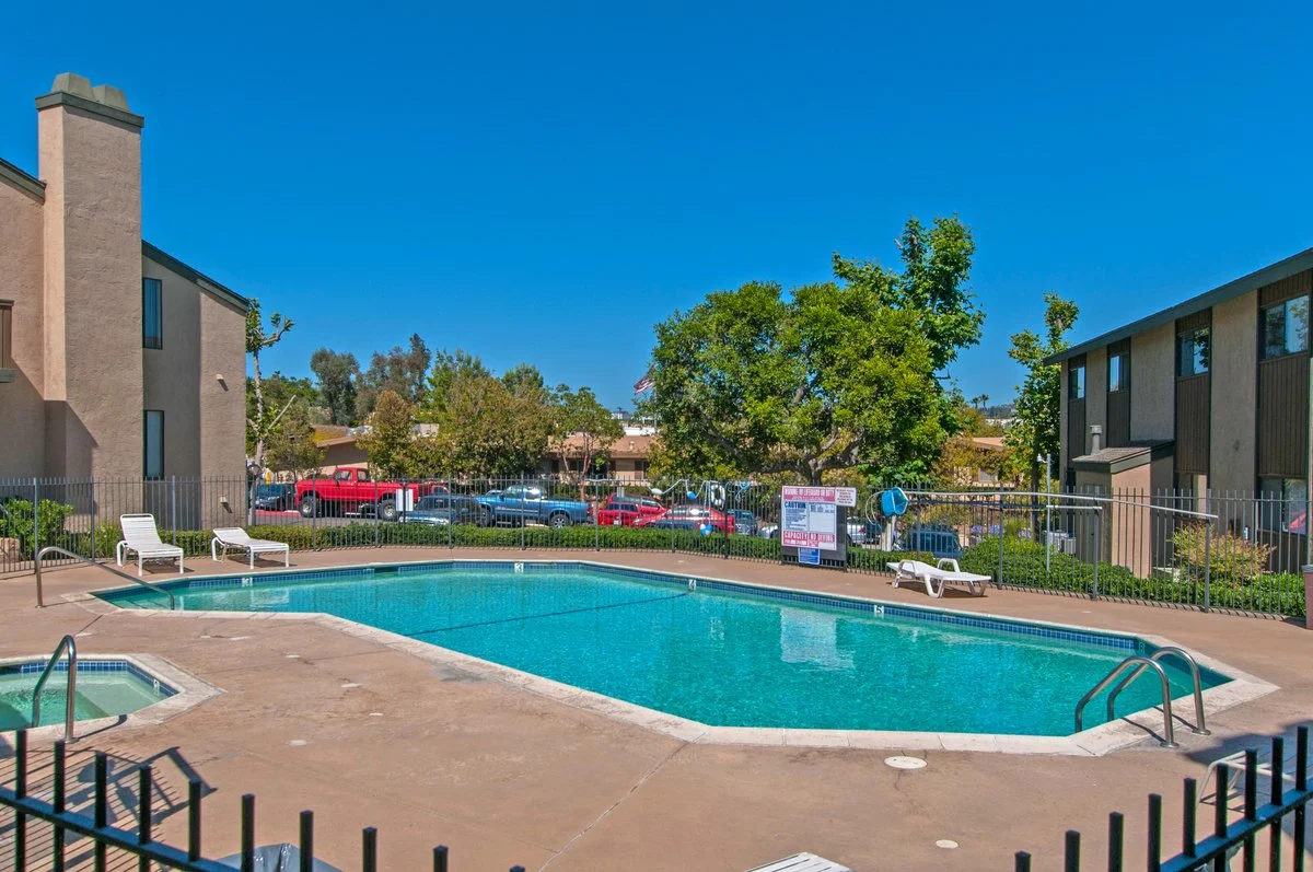 Apartment complex swimming pool with lounge chairs, surrounded by trees, with a parking lot and buildings in the background under a clear blue sky.