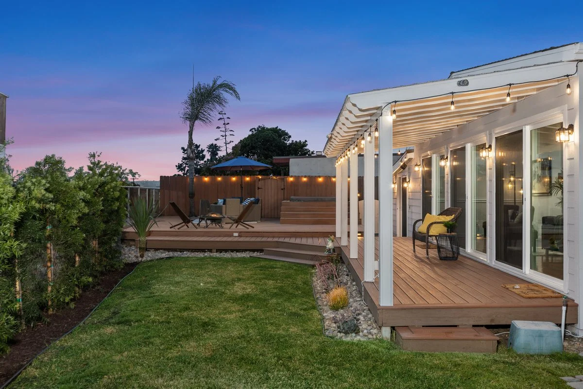Home backyard with wooden deck, outdoor seating area, string lights, grassy lawn, and trees at sunset.
