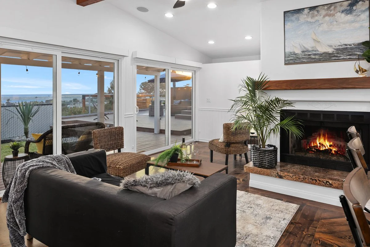 Living room with sliding glass doors leading to a deck with outdoor patio furniture, a cozy fireplace, a black sofa with throw pillows, a wooden coffee table with drinks, potted plants, and a view of the ocean outside.