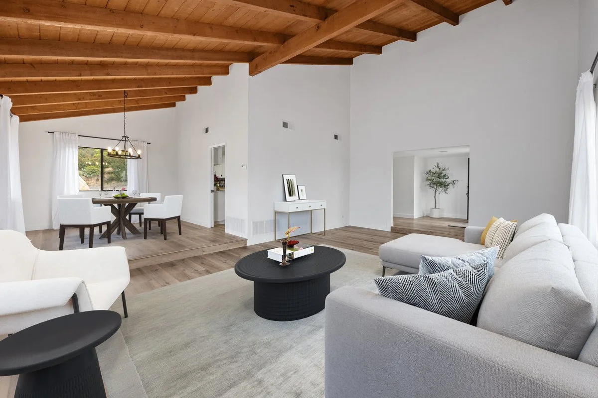 Living room with white sofa, black coffee table, and gray armchair, open to dining area with round wooden table and white chairs, wooden ceiling, white walls, large windows with white curtains, potted plant in an adjacent room.