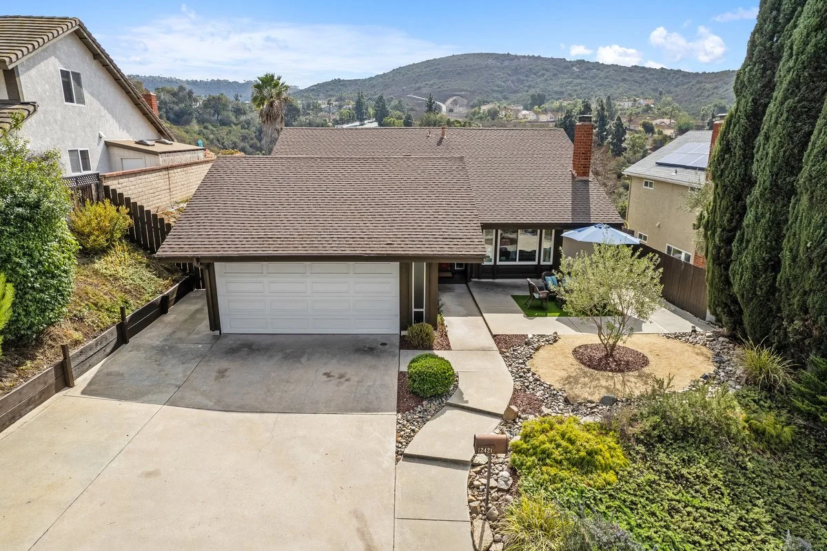 Aerial view of a suburban house with a brown roof, attached garage, small front yard with trees and shrubs, and a patio with outdoor furniture and umbrella, surrounded by neighboring homes and hills in the background.