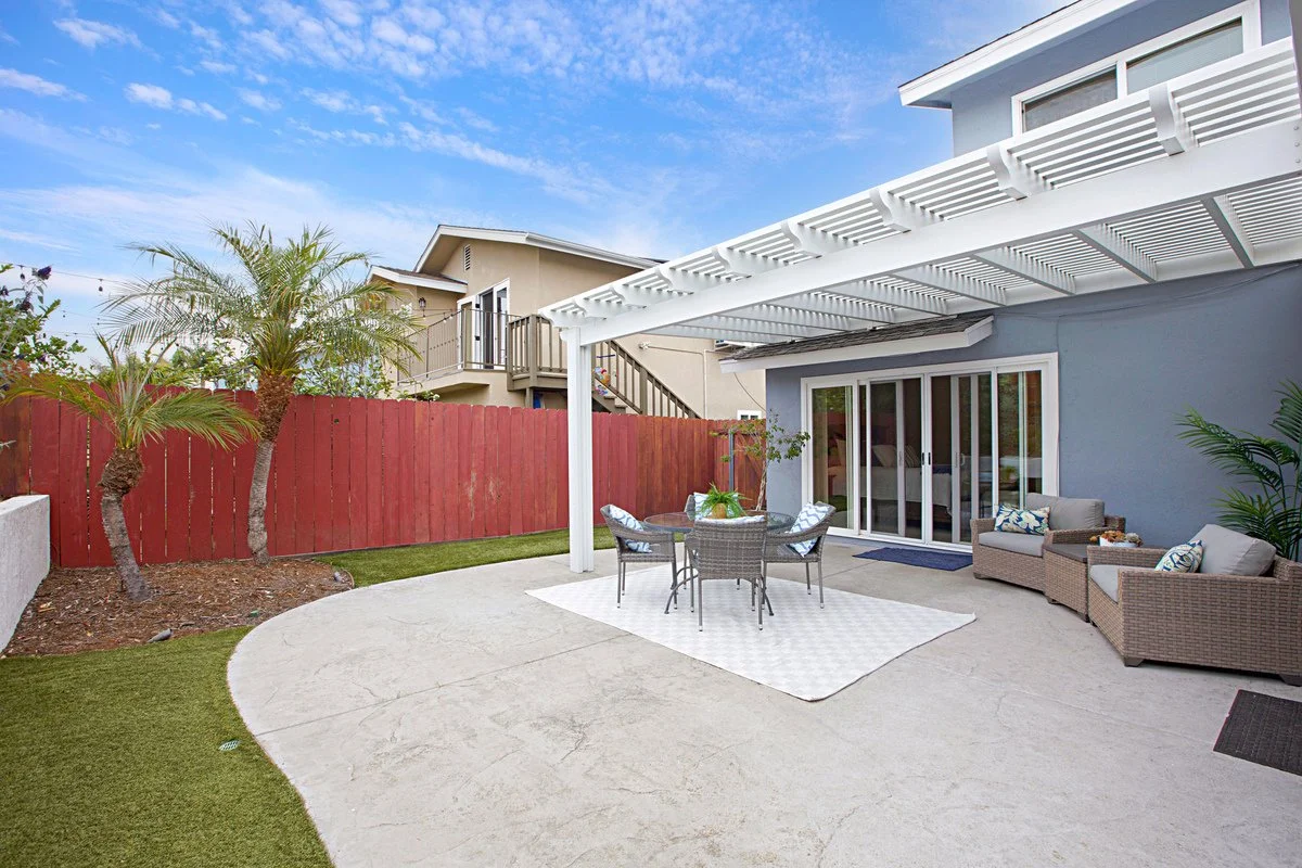 Backyard patio with outdoor furniture, including a round table with four chairs and a sitting area with a sofa and two armchairs, surrounded by a red wooden fence, potted plants, and a covered area with a white pergola, under a partly cloudy sky.