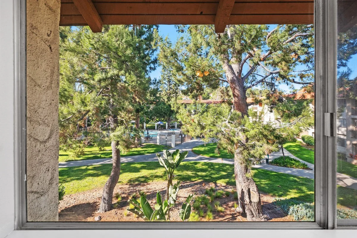View through a window showing trees, grass, and a park area with pathways and a pool with a fence around it.