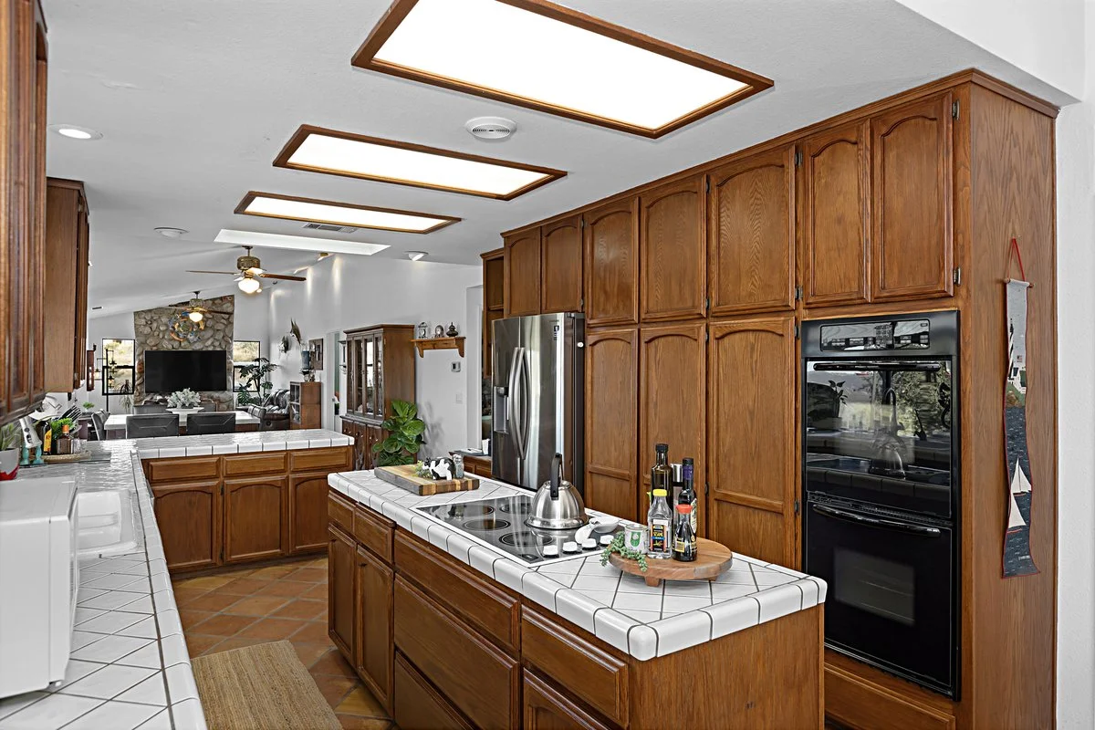 Kitchen with wooden cabinets, white tiled countertops, and a stove with a kettle. The room opens into a living room with a stone fireplace, a ceiling fan, and a large TV.
