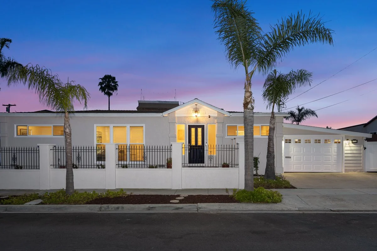 A modern white house with a black front door, surrounded by a white wall and black fence, palm trees, and a well-maintained yard at dusk.