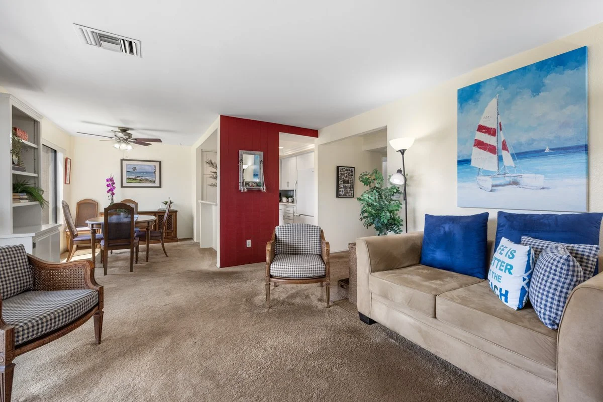 Living room with beige sofa, blue pillows, checkered armchair, wooden dining table with chairs, potted plant, wall art, and a red accent wall, with a doorway leading to the kitchen.