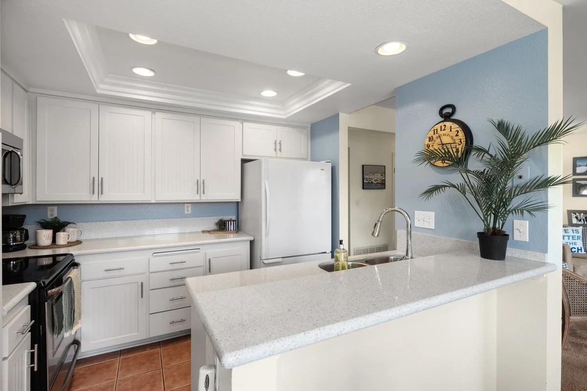 Modern kitchen with white cabinets, a white refrigerator, a countertop with a sink, and decorative items. Blue painted wall with a large wall clock, potted plant, and framed pictures in the adjacent room.
