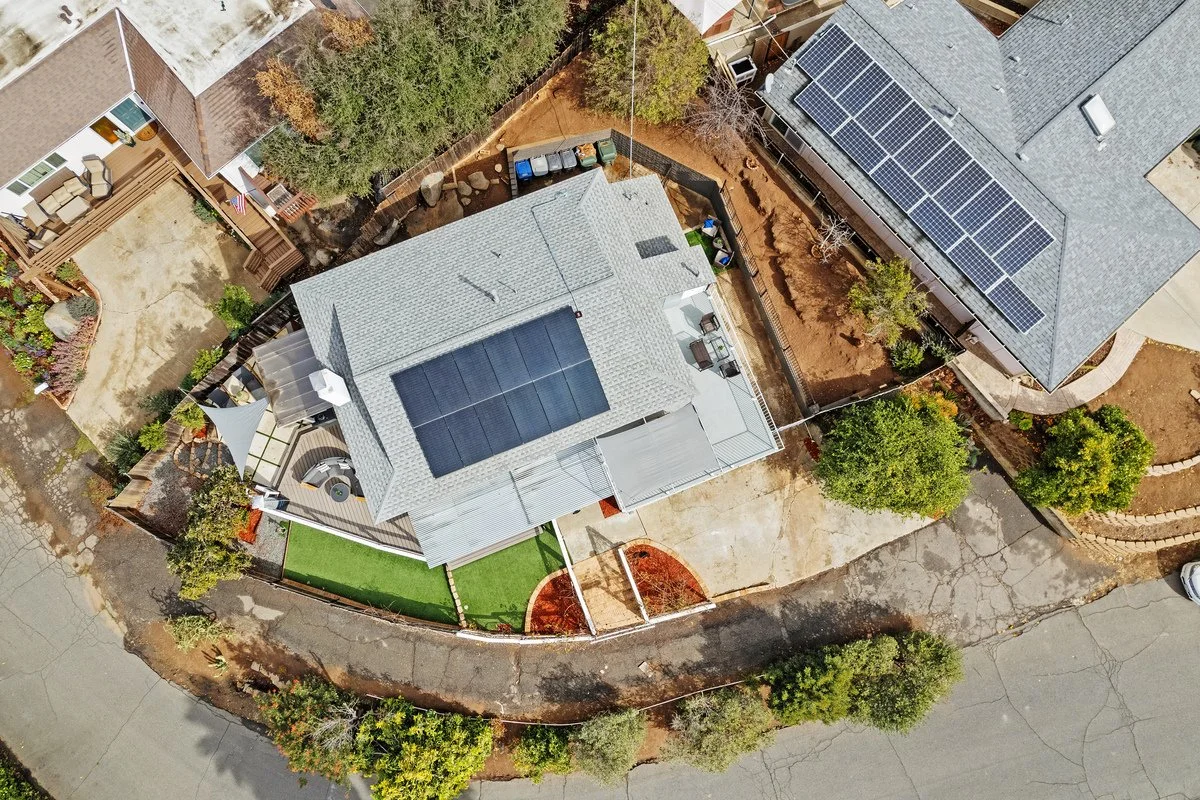 An aerial view of residential homes including a house with solar panels on the roof, fenced yard, outdoor furniture, and adjacent houses with solar panels.