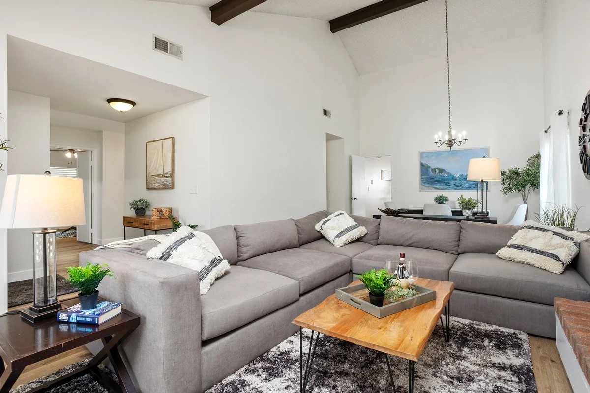 Living room with a large L-shaped gray sofa, a wooden coffee table with decorative items, and a black and white rug. The room features white walls, ceiling beams, and several lamps and framed art on the walls. There are houseplants near windows with 
