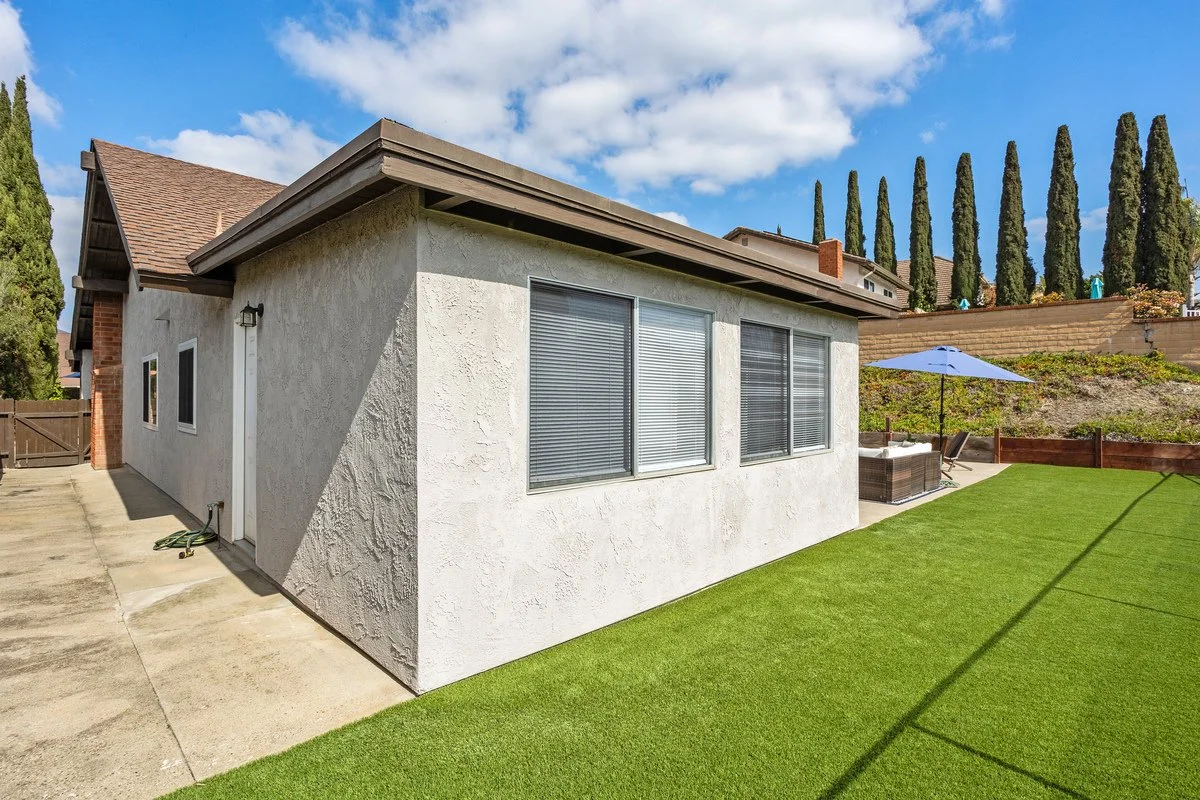 Backyard view of a single-story house with a white stucco exterior, three windows with blinds, a concrete patio, a grassy area, outdoor furniture with a blue umbrella, and a retaining wall with tall cypress trees behind it under a partly cloudy sky.