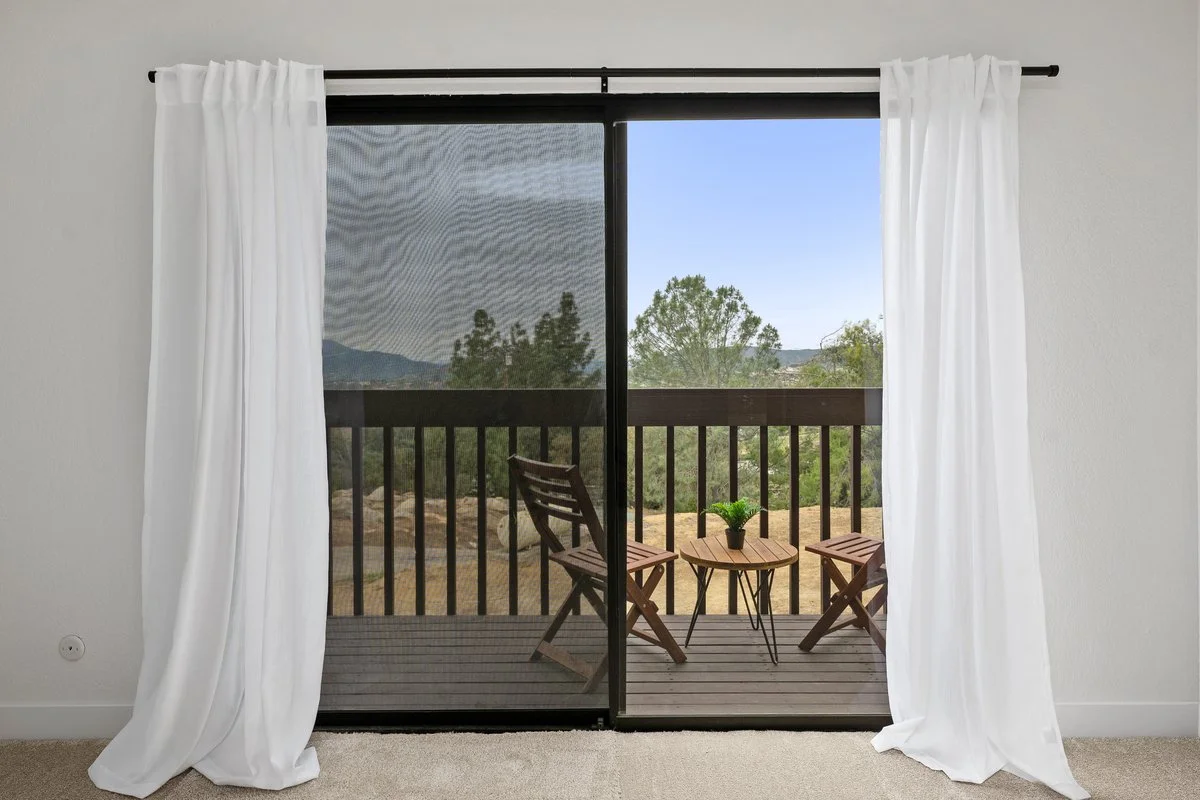 Sliding glass door with white curtains open to a balcony with two wooden chairs, a small table with a potted plant, and view of trees and mountains in the distance.