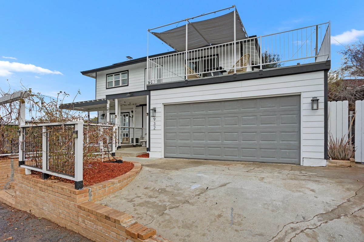 Two-story house with a white exterior, gray garage door, small front porch, and a large upstairs balcony with patio furniture and a shaded canopy. The driveway is concrete with cracks, and there is a brick and metal fence surrounding a small front ya