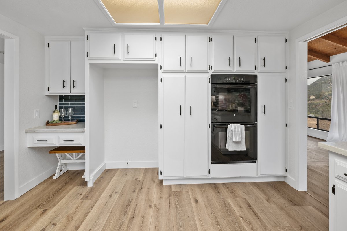 White kitchen cabinets with black hardware, double oven, and wood flooring, with a small breakfast nook.