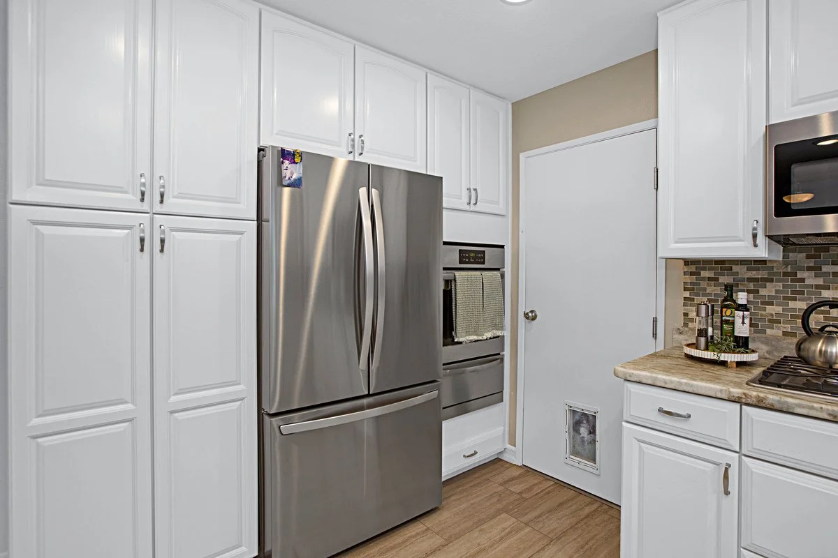 Kitchen with white cabinets, stainless steel refrigerator, microwave, oven, beige countertop, mosaic tile backsplash, and wood-look flooring.
