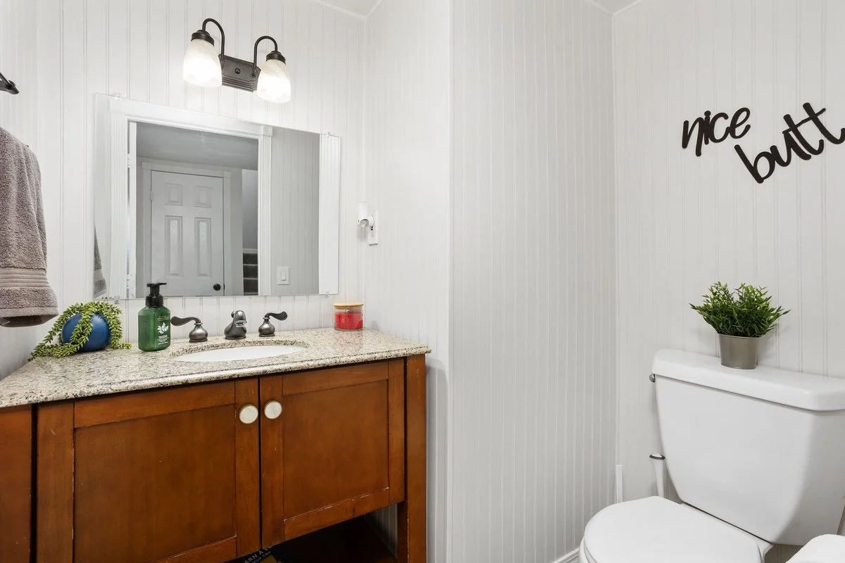 A bathroom with white paneled walls, a wooden vanity with a granite countertop, a mirror, and a toilet with a small potted plant on the tank. There is a towel hanging on the wall and a sign that says 'nice butt' on the wall.