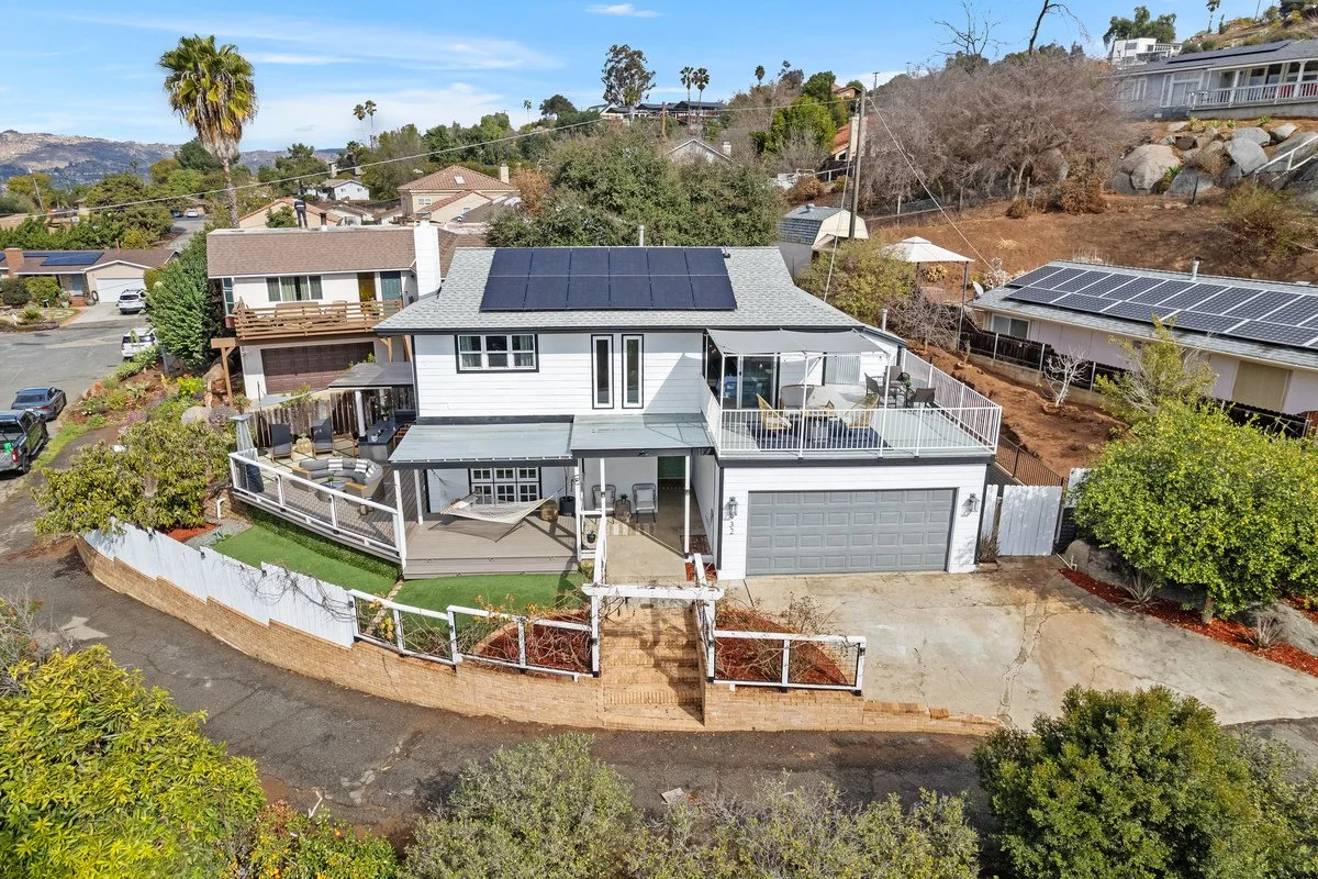 A two-story house with solar panels on the roof, surrounded by a fenced yard with a deck and patio, located in a suburban neighborhood with other houses and trees.