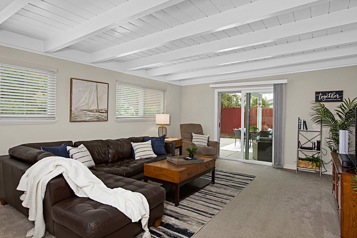 Living room with a black leather sectional sofa with white and navy throw pillows, a wooden coffee table, beige armchair, large windows with blinds, sliding glass door leading to an outdoor patio, and a bookshelf with decorations and a plant.