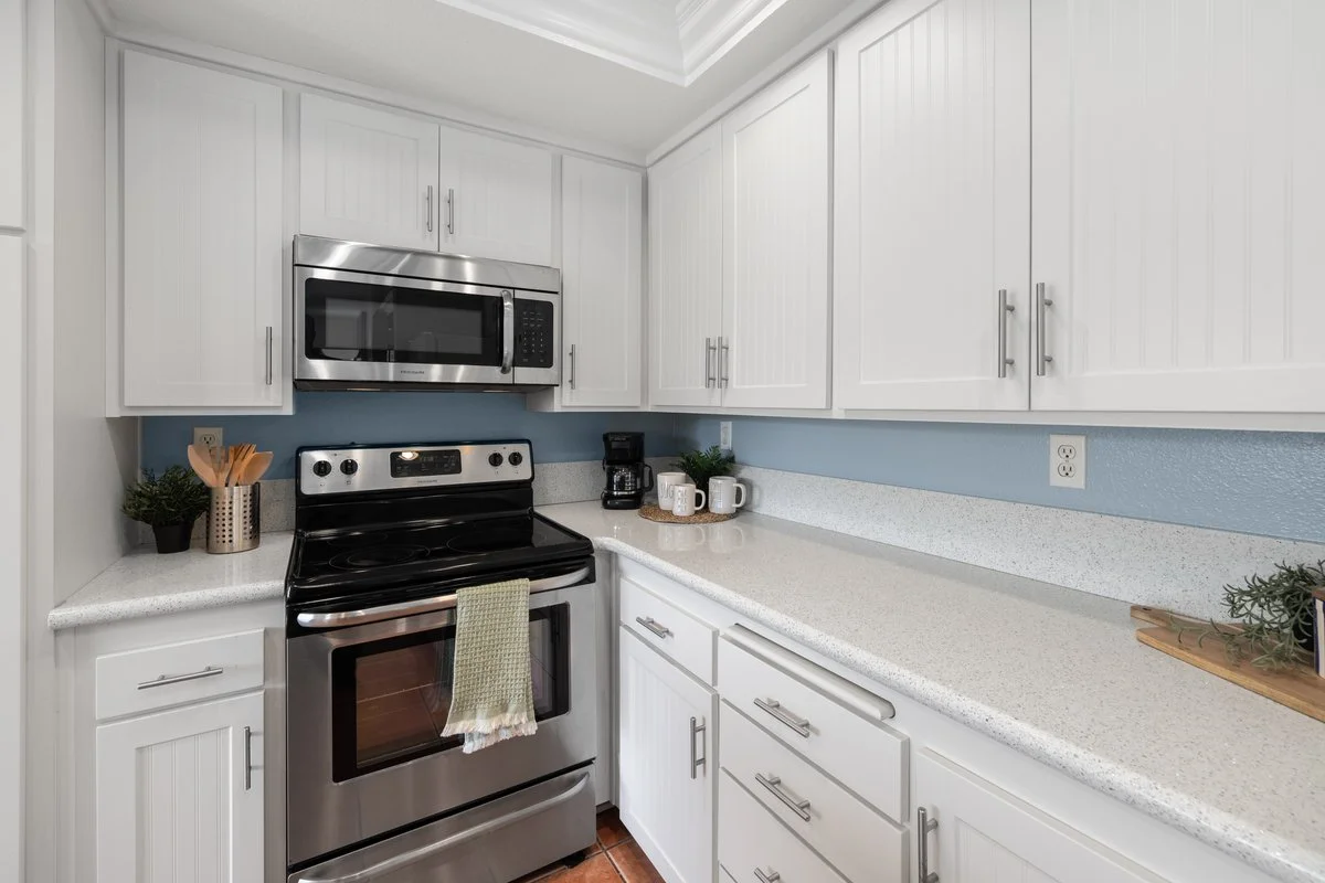 Modern kitchen with white cabinets, stainless steel microwave and stove, counter with coffee maker, mugs, and small plants.