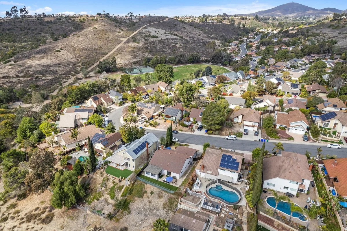 Aerial view of a residential neighborhood situated near a hilly area with dry terrain and few trees, featuring houses with solar panels, some with swimming pools, and a school yard with sports courts. In the background, hills and mountains are visibl