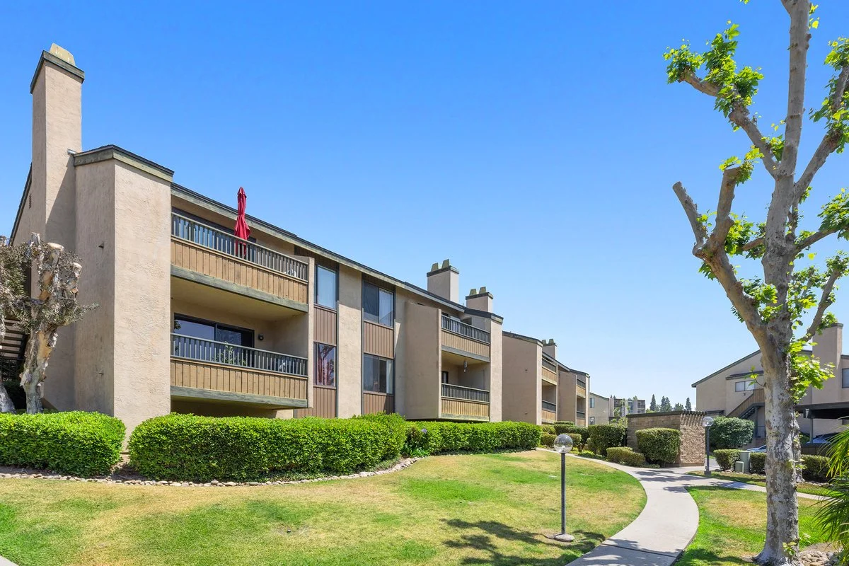 Exterior view of a multi-story apartment complex with balconies, surrounded by well-maintained green lawns, trees, and bushes, under a clear blue sky.