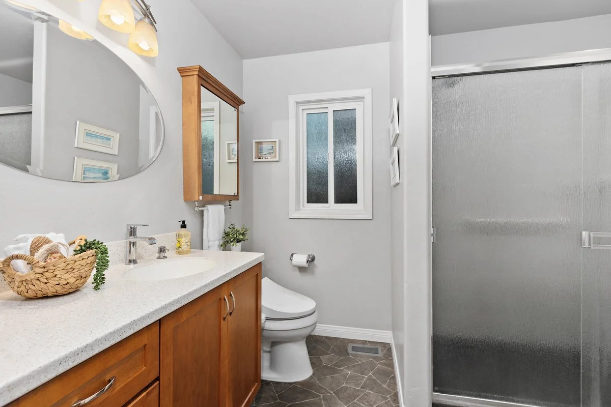A bathroom with a wooden vanity, a white countertop, a mirror, a toilet, a frosted window, and a glass shower door.