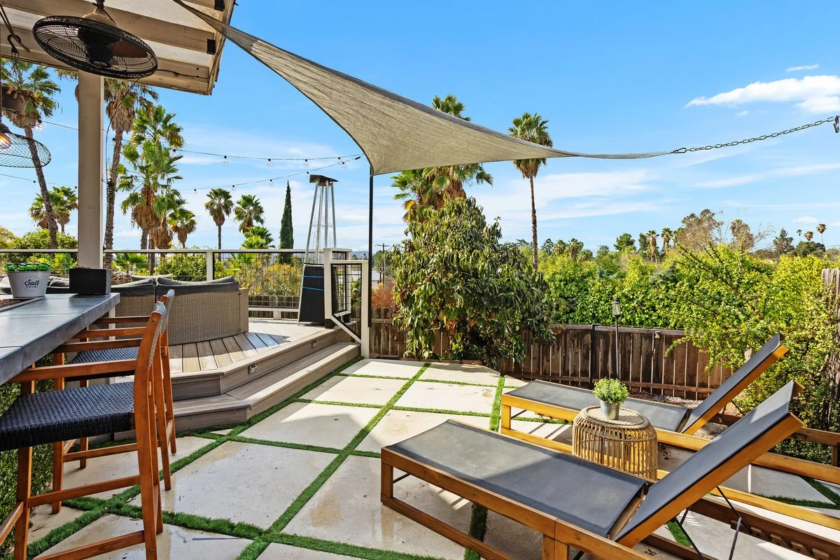 Outdoor patio with lounge chairs, a small table with a plant, a deck area with garden furniture, string lights, and lush trees and palm trees in a sunny sky.