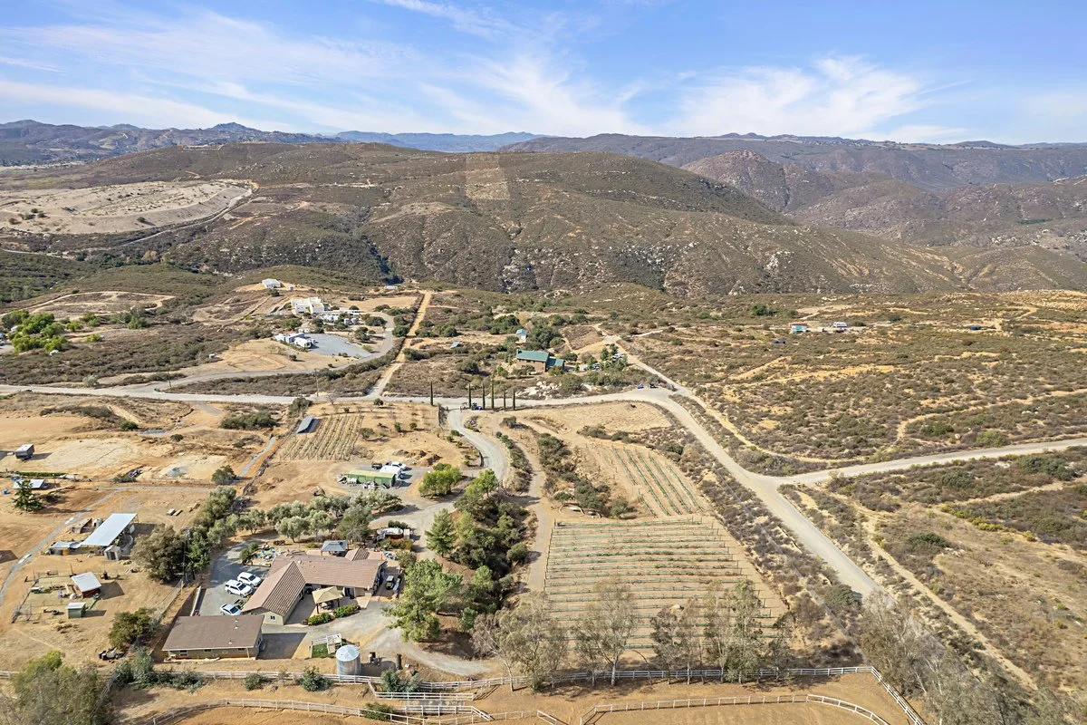 Aerial view of a rural landscape with houses, dirt roads, farmland, and hills in the background under a partly cloudy sky.