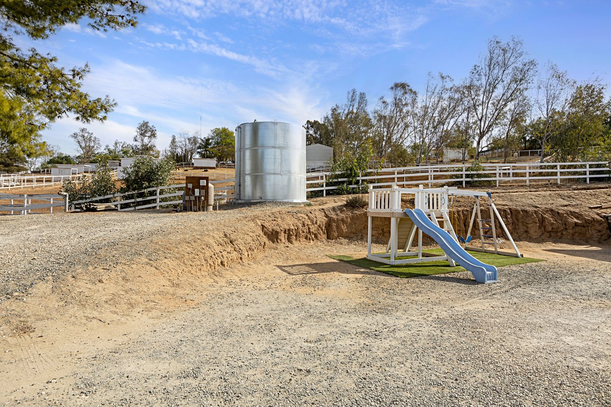 A small playground with a slide and swings situated on a grassy patch in a dirt area. In the background, there is a water tank, trees, and a white fenced area on a sunny day with a blue sky.