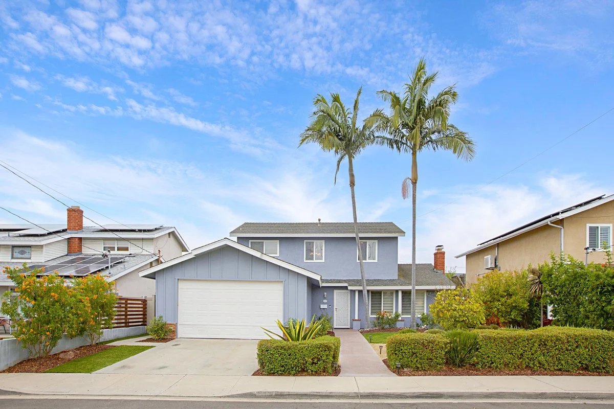 A suburban house with a blue exterior, two-story structure, white garage door, and landscaped front yard with palm trees and bushes.