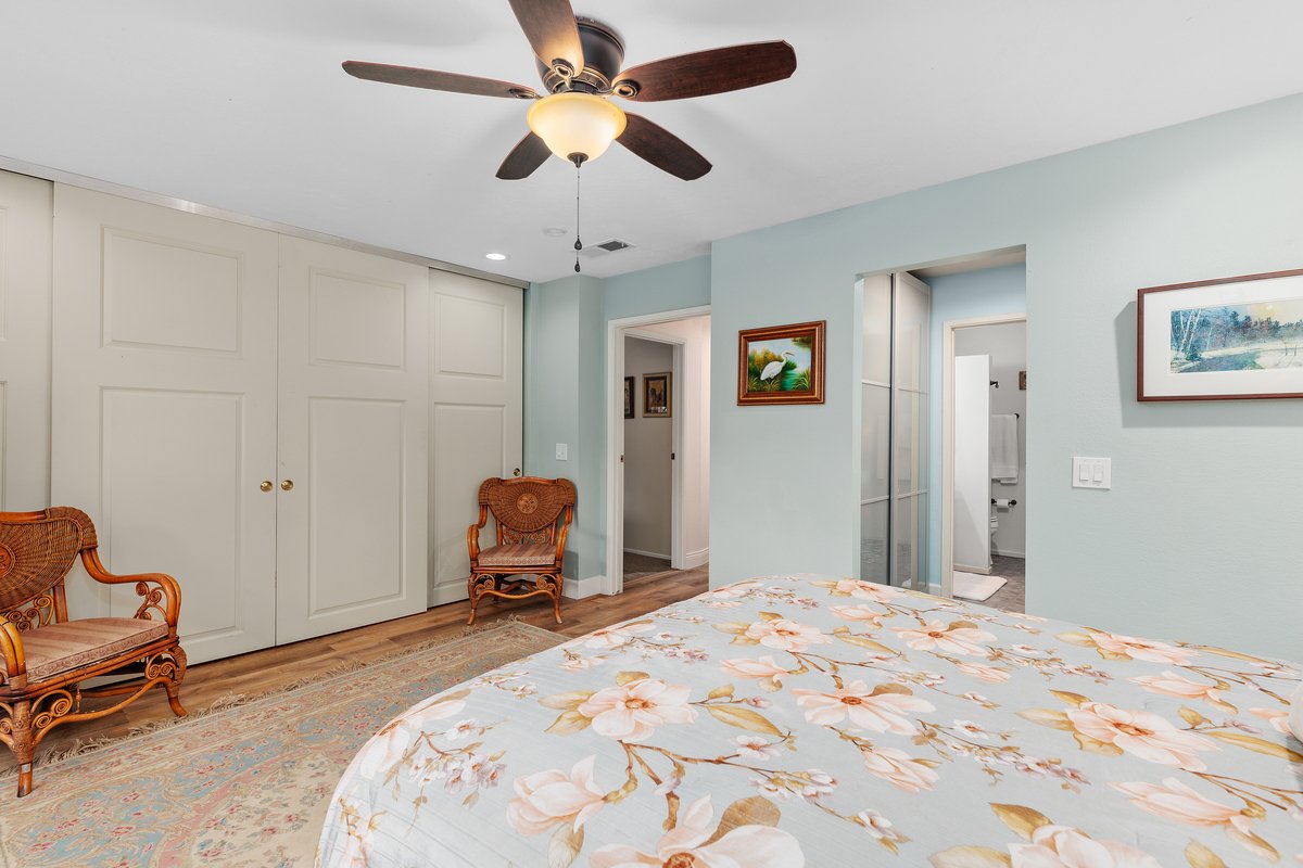 A bedroom with a bed featuring a floral bedspread, two wooden chairs with woven seats, a ceiling fan with wooden blades, and a closet with closed white doors. There are framed pictures on the walls and an open doorway leading to a bathroom.