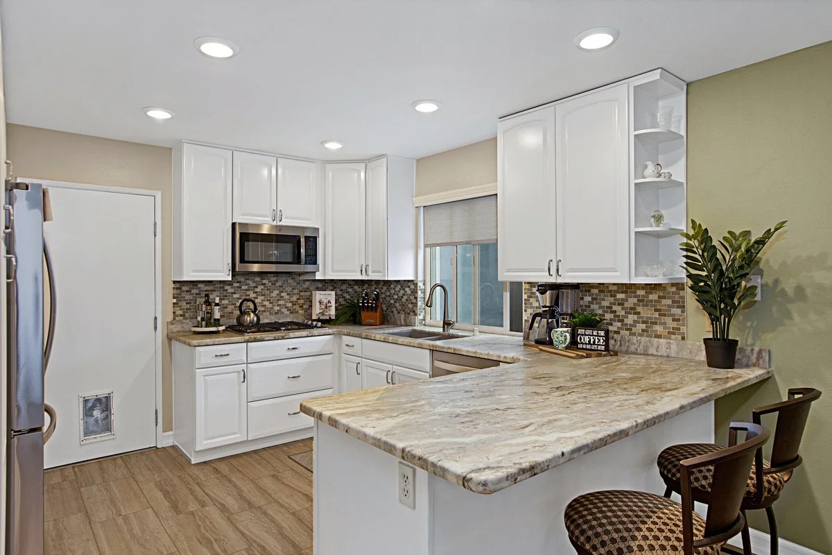 Kitchen with white cabinets, granite countertops, mosaic tile backsplash, stainless steel microwave, and refrigerator. There is a small breakfast bar with two chairs, a window with a beige shade, and some decorative items including a plant and coffee